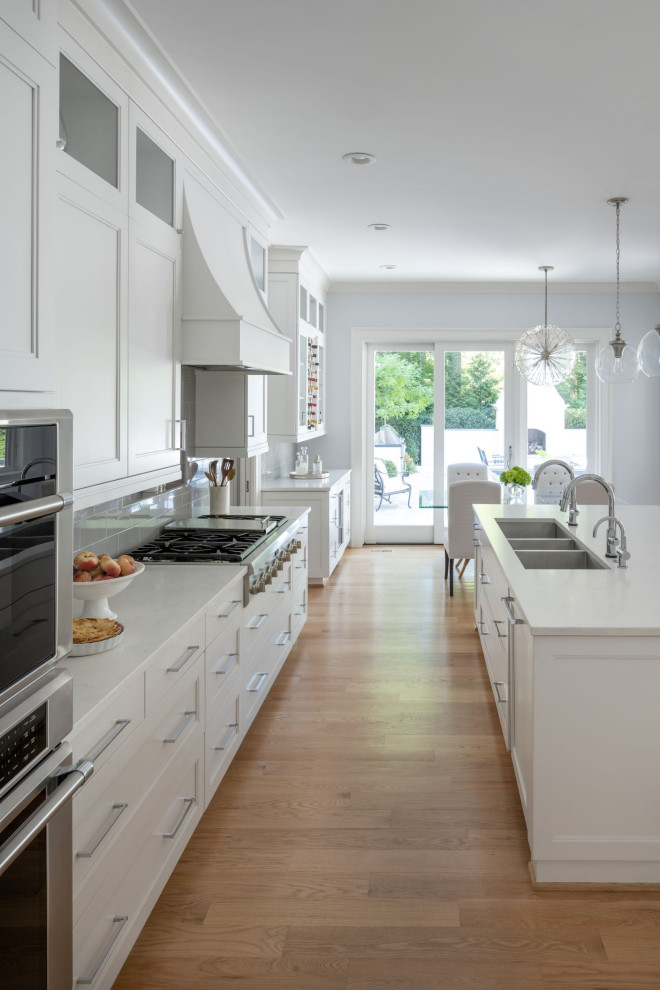 Photo of an expansive u-shaped eat-in kitchen in DC Metro with a triple-bowl sink, flat-panel cabinets, white cabinets, quartzite benchtops, grey splashback, subway tile splashback, panelled appliances, medium hardwood floors, with island, brown floor and white benchtop.