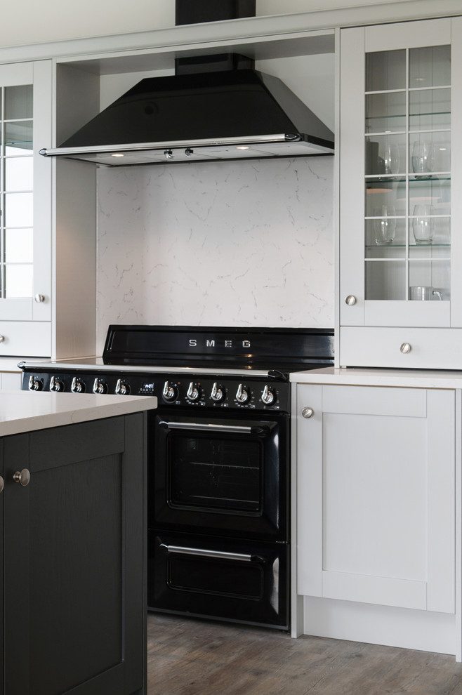 Large traditional l-shaped eat-in kitchen in Devon with a farmhouse sink, shaker cabinets, grey cabinets, quartzite benchtops, white splashback, glass sheet splashback, black appliances, laminate floors, with island, brown floor and white benchtop.