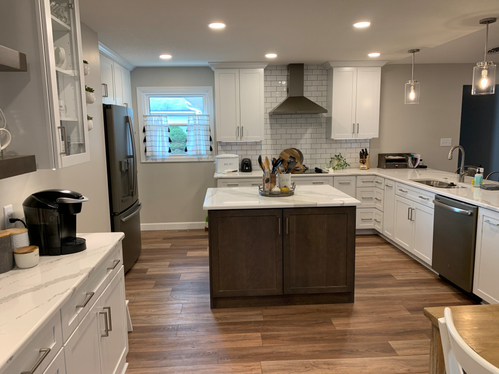 Large transitional u-shaped vinyl floor and brown floor open concept kitchen photo with an undermount sink, shaker cabinets, white cabinets, quartz countertops, white backsplash, ceramic backsplash, stainless steel appliances, an island and white countertops