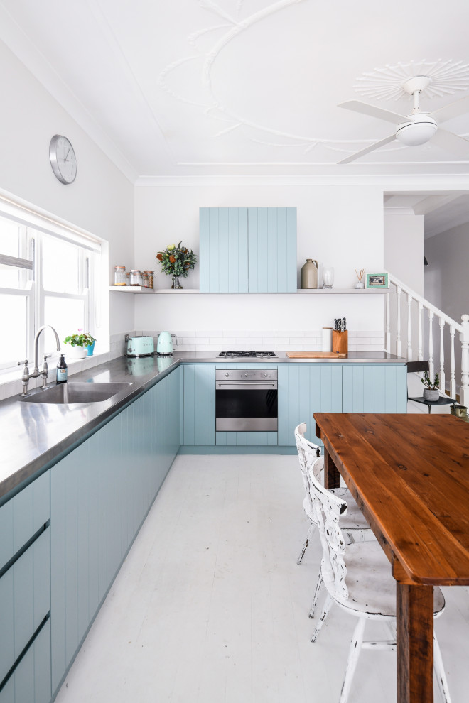 Design ideas for a small beach style l-shaped eat-in kitchen in Sydney with an undermount sink, blue cabinets, stainless steel benchtops, white splashback, subway tile splashback, stainless steel appliances, light hardwood floors, no island, white floor and grey benchtop.