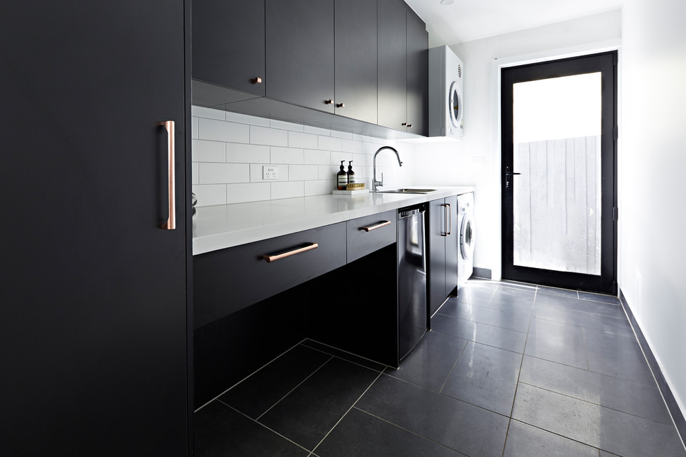 Photo of a mid-sized modern single-wall dedicated laundry room in Melbourne with an undermount sink, black cabinets, a stacked washer and dryer, black floor, white benchtop, quartz benchtops and white walls.