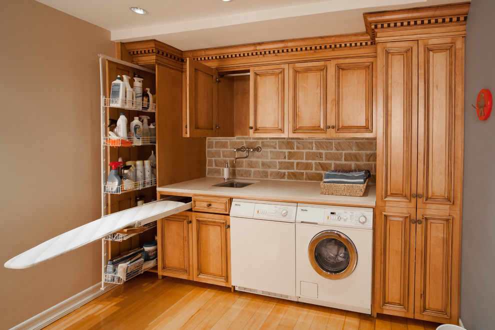 This is an example of a small traditional single-wall utility room in New York with a single-bowl sink, raised-panel cabinets, light hardwood floors, a side-by-side washer and dryer, medium wood cabinets, granite benchtops, multi-coloured walls, brown floor and beige benchtop.