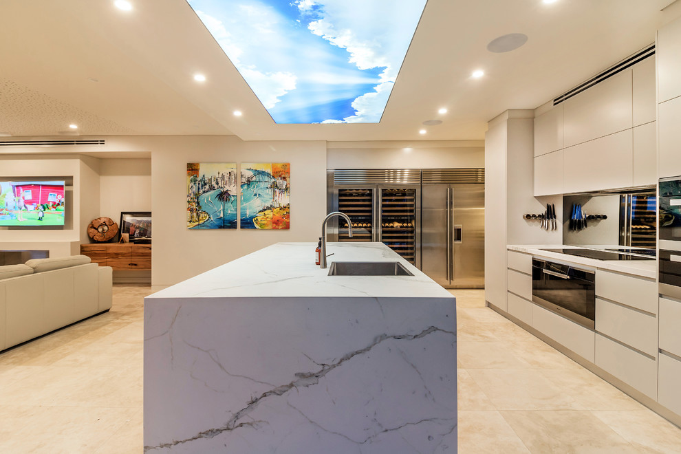 This is an example of a contemporary open plan kitchen in Sydney with a single-bowl sink, flat-panel cabinets, beige cabinets, metallic splashback, mirror splashback, black appliances, with island, beige floor and beige benchtop.
