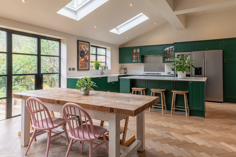Photo of a large classic l-shaped open plan kitchen in London with a belfast sink, shaker cabinets, green cabinets, quartz worktops, metallic splashback, integrated appliances, medium hardwood flooring, an island and white worktops.