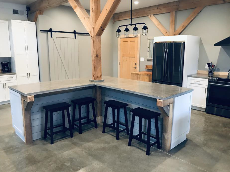 Inspiration for an expansive country l-shaped eat-in kitchen in Chicago with a farmhouse sink, shaker cabinets, white cabinets, concrete benchtops, timber splashback, black appliances, concrete floors, with island, grey floor, grey benchtop and exposed beam.