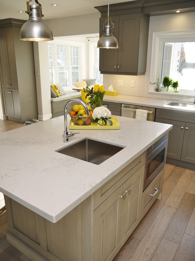 Example of a mid-sized transitional l-shaped light wood floor enclosed kitchen design in Toronto with an undermount sink, recessed-panel cabinets, gray cabinets, quartz countertops, white backsplash, porcelain backsplash, stainless steel appliances and an island