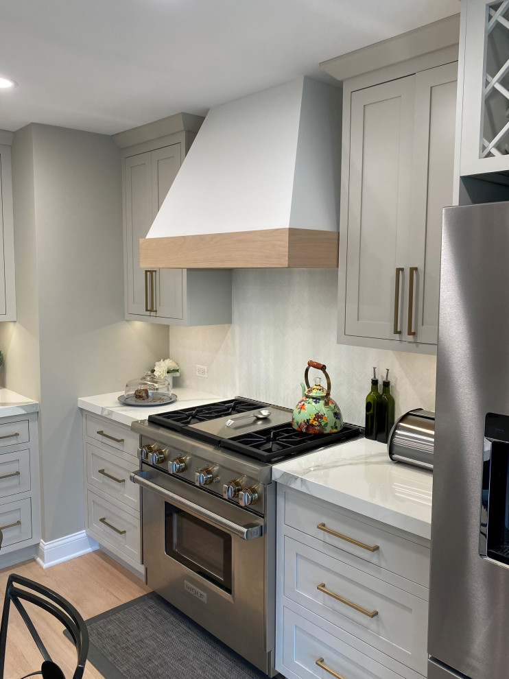 Design ideas for a medium sized classic l-shaped kitchen/diner in Chicago with a belfast sink, flat-panel cabinets, grey cabinets, white splashback, stone tiled splashback, stainless steel appliances, medium hardwood flooring, an island, brown floors and white worktops.