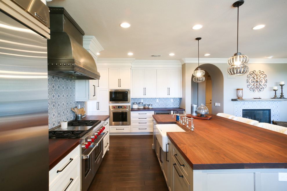 This is an example of a large modern l-shaped eat-in kitchen in San Diego with a farmhouse sink, shaker cabinets, white cabinets, wood benchtops, blue splashback, cement tile splashback, stainless steel appliances, dark hardwood floors, with island, brown floor and brown benchtop.