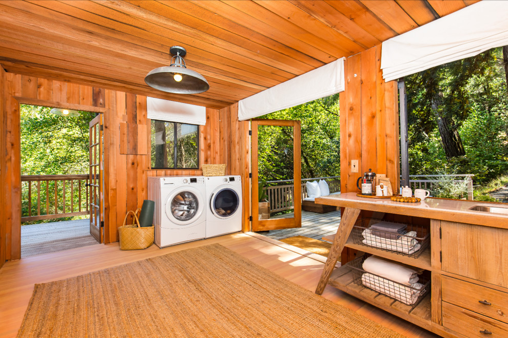Photo of a large country utility room in San Francisco with light hardwood floors, a side-by-side washer and dryer, open cabinets, medium wood cabinets and orange floor.