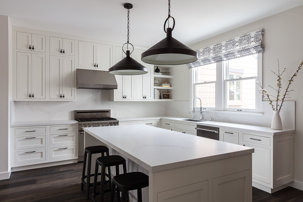 Example of a transitional l-shaped dark wood floor and brown floor kitchen design with an undermount sink, shaker cabinets, white cabinets, white backsplash, stone slab backsplash, stainless steel appliances, an island and white countertops