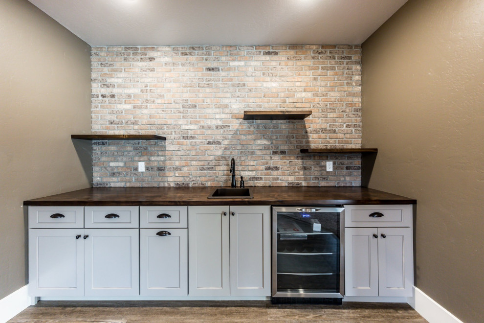 Mid-sized transitional single-wall wet bar in St Louis with a drop-in sink, recessed-panel cabinets, white cabinets, wood benchtops, beige splashback, brick splashback, dark hardwood floors, brown floor and brown benchtop.