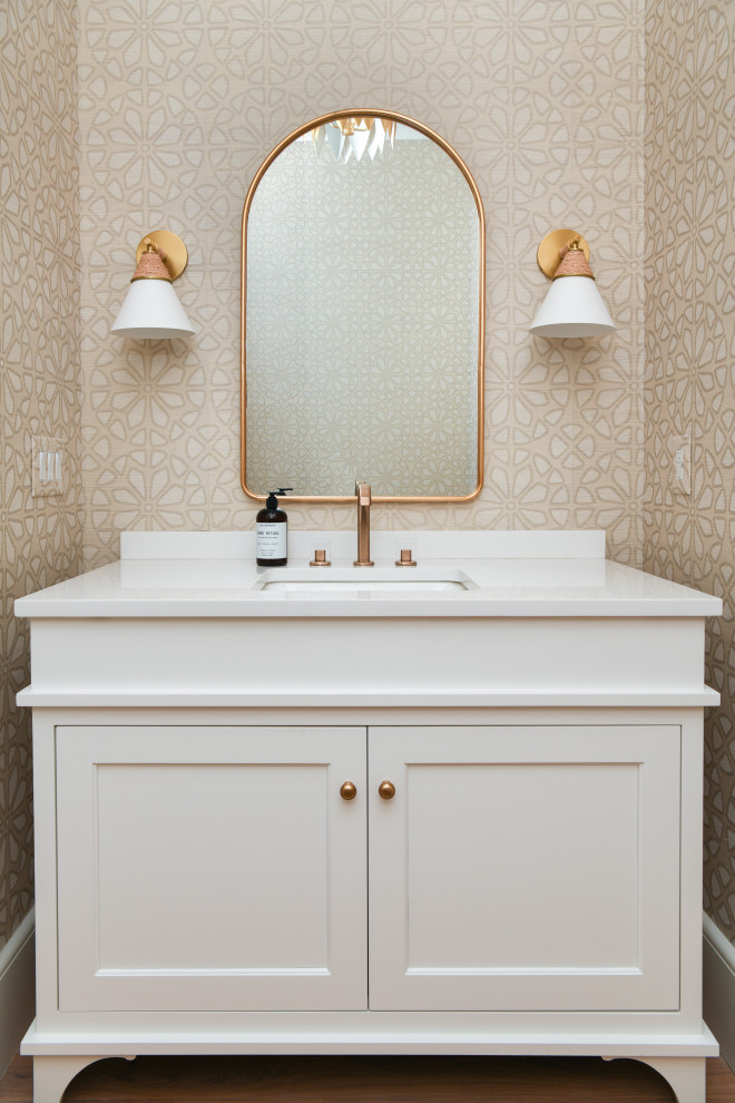 Example of a beach style medium tone wood floor, brown floor and wallpaper powder room design in Charlotte with shaker cabinets, white cabinets, beige walls, an undermount sink, white countertops and a freestanding vanity