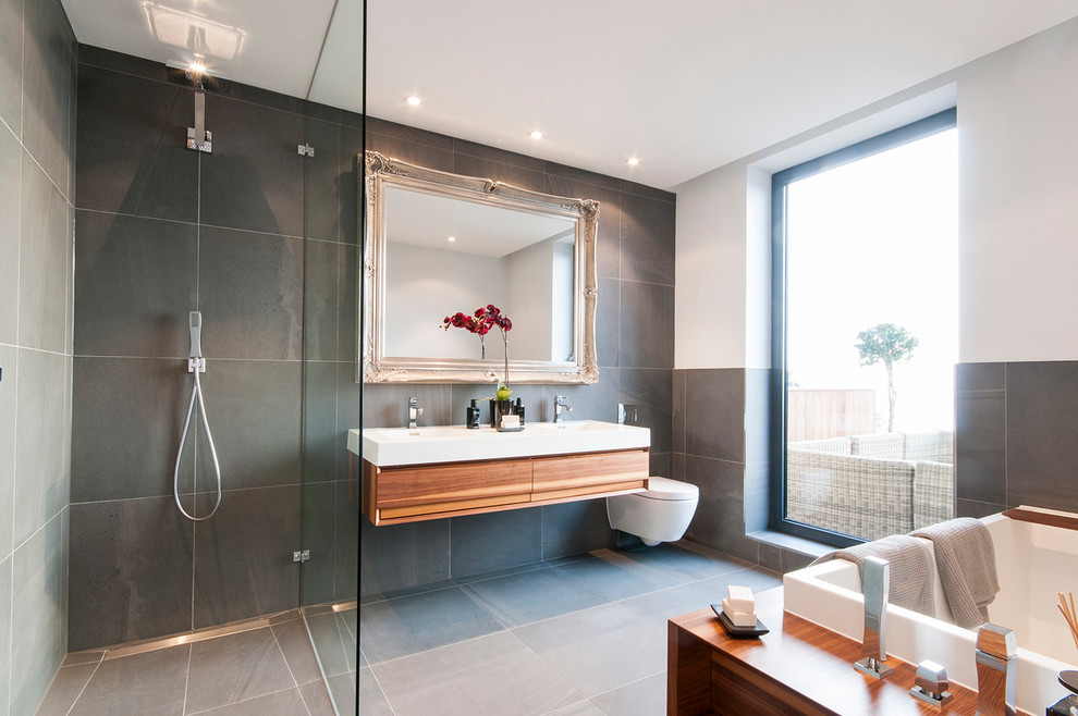 Mid-sized contemporary bathroom in London with a wall-mount toilet, gray tile, a wall-mount sink, flat-panel cabinets, medium wood cabinets, white walls and a curbless shower.