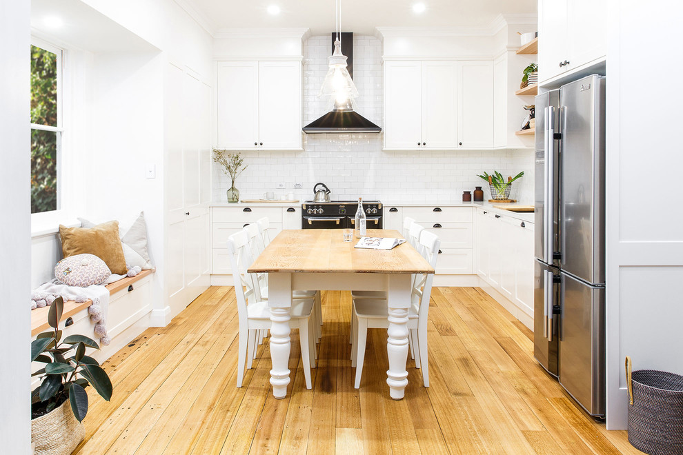 Photo of a traditional u-shaped eat-in kitchen in Adelaide with shaker cabinets, white cabinets, white splashback, subway tile splashback, black appliances and light hardwood floors.