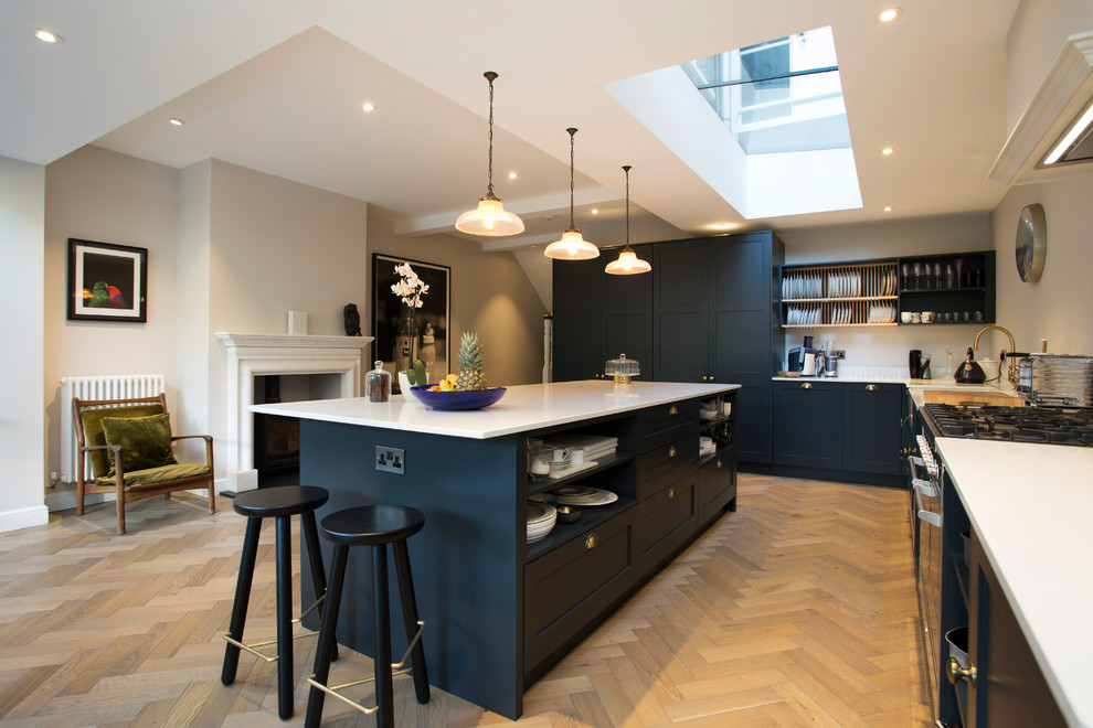 This is an example of a large transitional l-shaped kitchen in London with shaker cabinets, blue cabinets, quartzite benchtops, light hardwood floors, with island and white benchtop.