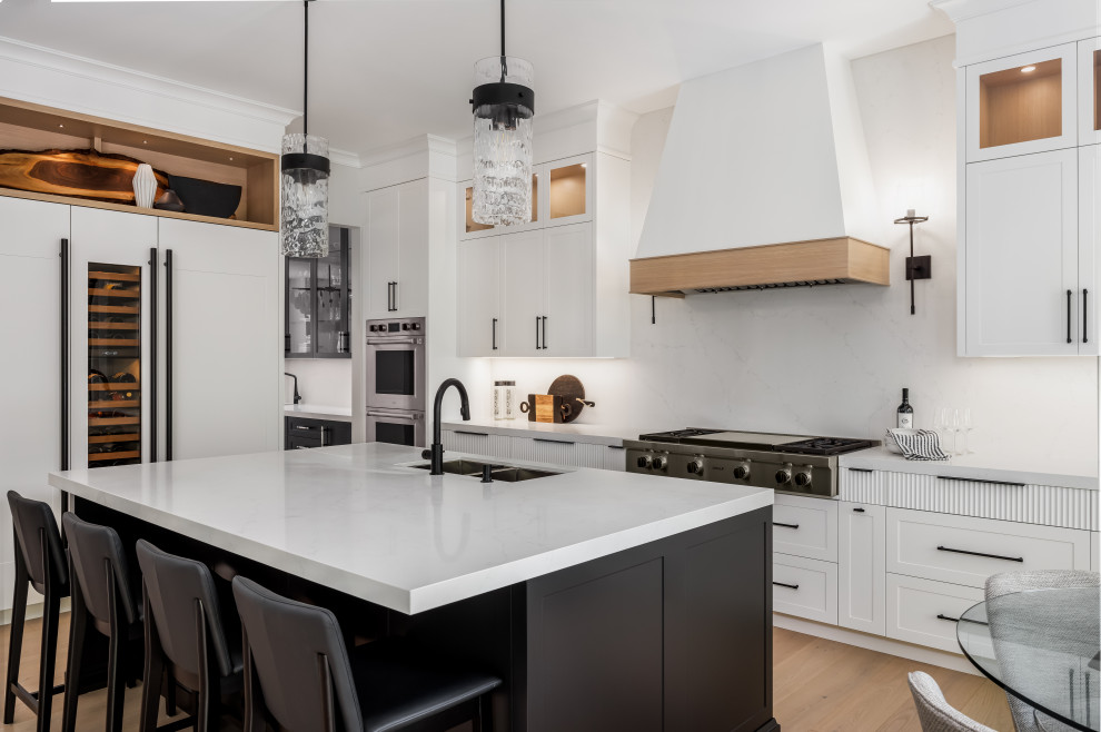 Example of a transitional l-shaped medium tone wood floor and brown floor eat-in kitchen design in Toronto with an undermount sink, shaker cabinets, white cabinets, white backsplash, stone slab backsplash, stainless steel appliances, an island and white countertops