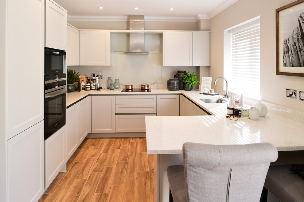 This is an example of a mid-sized transitional u-shaped separate kitchen in London with porcelain floors, brown floor, an undermount sink, recessed-panel cabinets, grey cabinets, glass sheet splashback, a peninsula, white benchtop, beige splashback and panelled appliances.