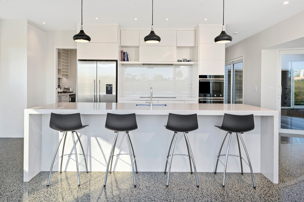 This is an example of a large modern single-wall eat-in kitchen in Auckland with a drop-in sink, raised-panel cabinets, white cabinets, white splashback, glass sheet splashback, stainless steel appliances, concrete floors, with island, multi-coloured floor and white benchtop.