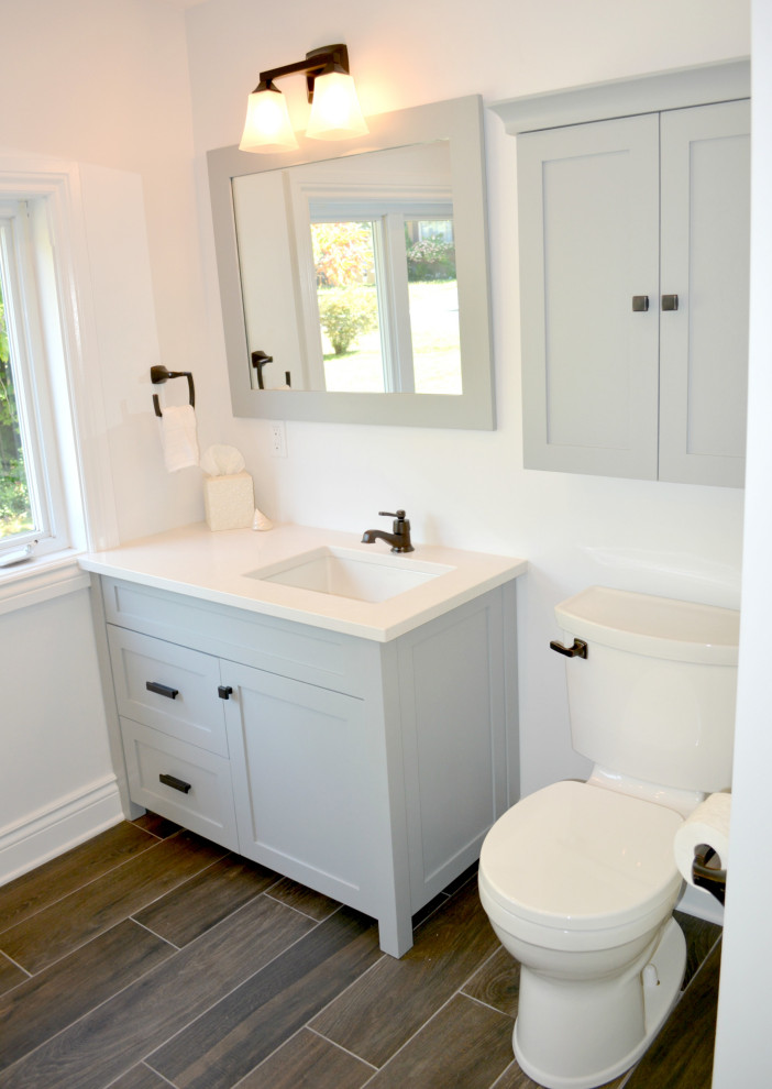 Cette image montre une petite salle de bain chalet avec un placard à porte shaker, des portes de placard grises, une baignoire en alcôve, un combiné douche/baignoire, WC séparés, un carrelage gris, des carreaux de céramique, un mur gris, un sol en carrelage de céramique, un lavabo encastré, un plan de toilette en quartz modifié, un sol marron, une cabine de douche à porte coulissante, un plan de toilette blanc, buanderie et meuble simple vasque.