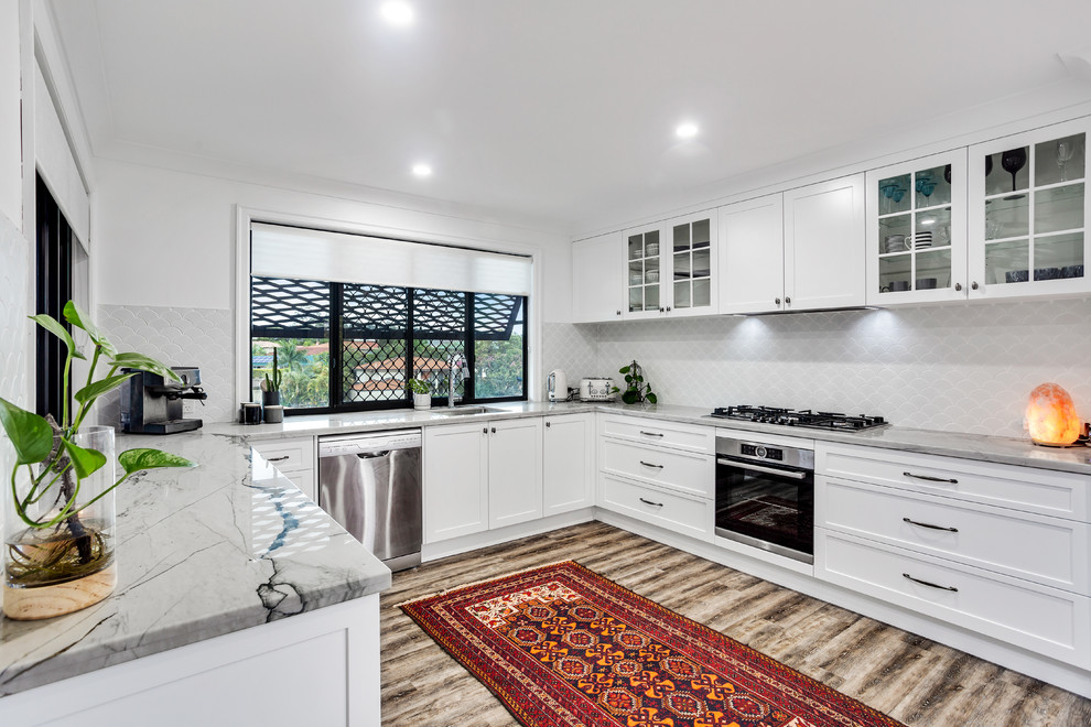 This is an example of a large transitional u-shaped eat-in kitchen in Sunshine Coast with an undermount sink, shaker cabinets, white cabinets, quartzite benchtops, white splashback, ceramic splashback, stainless steel appliances, vinyl floors, with island and white benchtop.