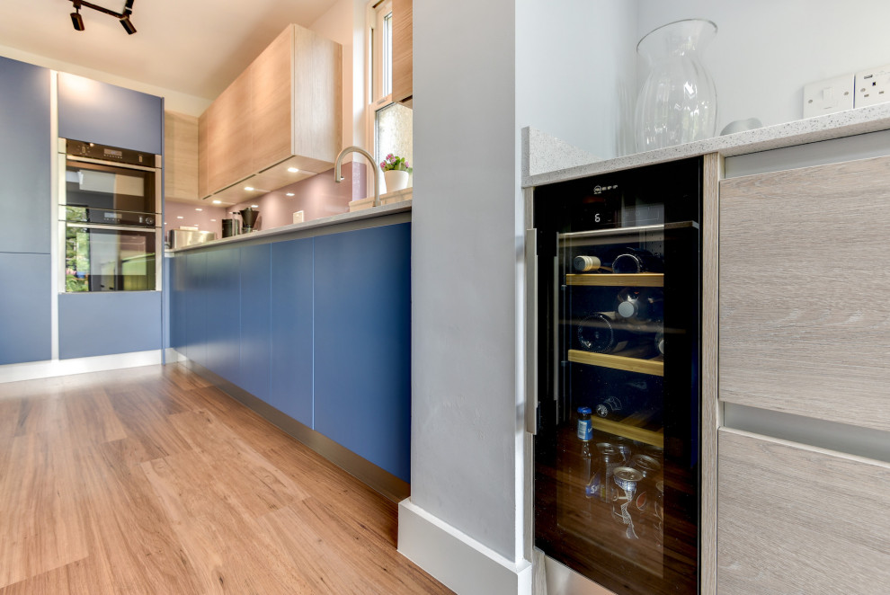 Inspiration for a medium sized nautical l-shaped kitchen/diner in Sussex with a double-bowl sink, flat-panel cabinets, blue cabinets, quartz worktops, multi-coloured splashback, glass sheet splashback, black appliances, dark hardwood flooring, an island, brown floors, white worktops and feature lighting.