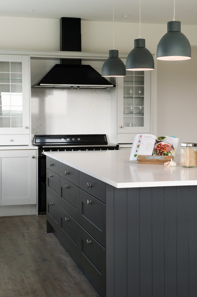 This is an example of a large traditional l-shaped eat-in kitchen in Devon with a farmhouse sink, shaker cabinets, grey cabinets, quartzite benchtops, white splashback, glass sheet splashback, black appliances, laminate floors, with island, brown floor and white benchtop.