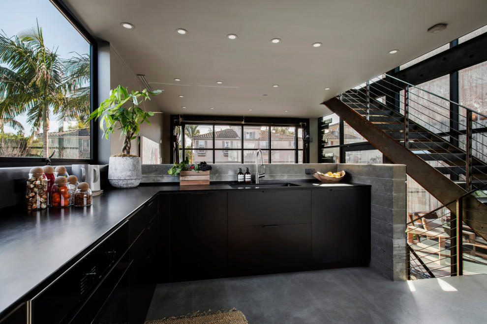 Photo of a mid-sized industrial l-shaped kitchen in Los Angeles with a single-bowl sink, flat-panel cabinets, black cabinets, grey splashback, stainless steel appliances, no island, grey floor and black benchtop.