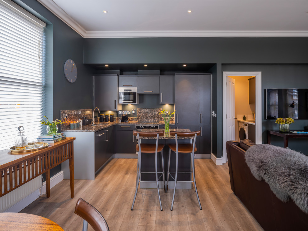 Photo of a mid-sized contemporary l-shaped open plan kitchen in Surrey with a drop-in sink, flat-panel cabinets, grey cabinets, granite benchtops, granite splashback, stainless steel appliances, with island, brown floor, multi-coloured splashback, laminate floors and multi-coloured benchtop.