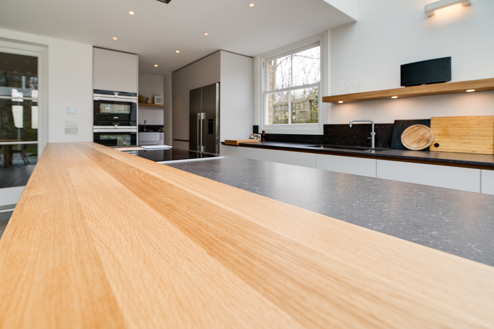 Large contemporary galley open plan kitchen in London with an undermount sink, flat-panel cabinets, white cabinets, solid surface benchtops, grey splashback, stainless steel appliances, ceramic floors, with island, grey floor and grey benchtop.