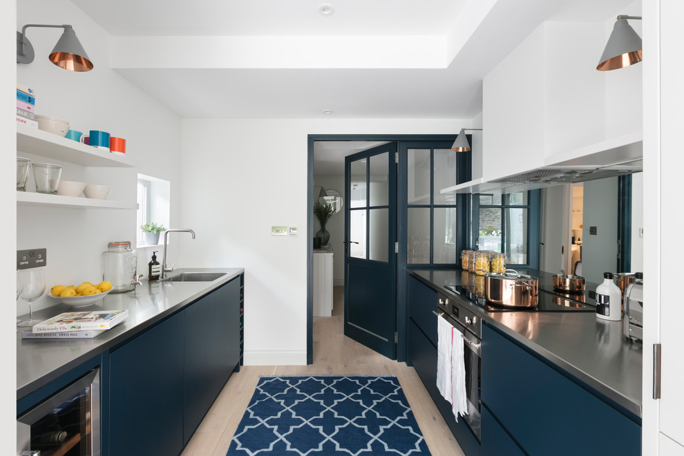 Inspiration for a mid-sized scandinavian galley eat-in kitchen in London with a single-bowl sink, blue cabinets, stainless steel benchtops, metallic splashback, mirror splashback, panelled appliances, light hardwood floors and glass-front cabinets.