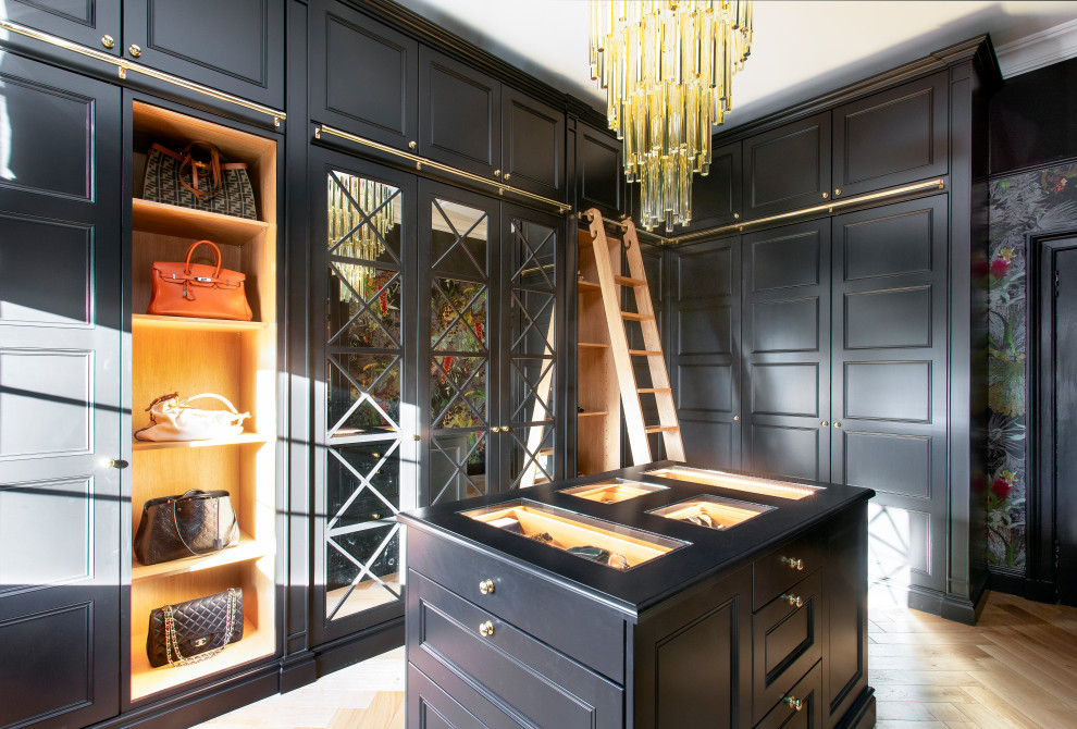 This is an example of a large classic gender neutral walk-in wardrobe in Edinburgh with glass-front cabinets, black cabinets, light hardwood flooring and beige floors.