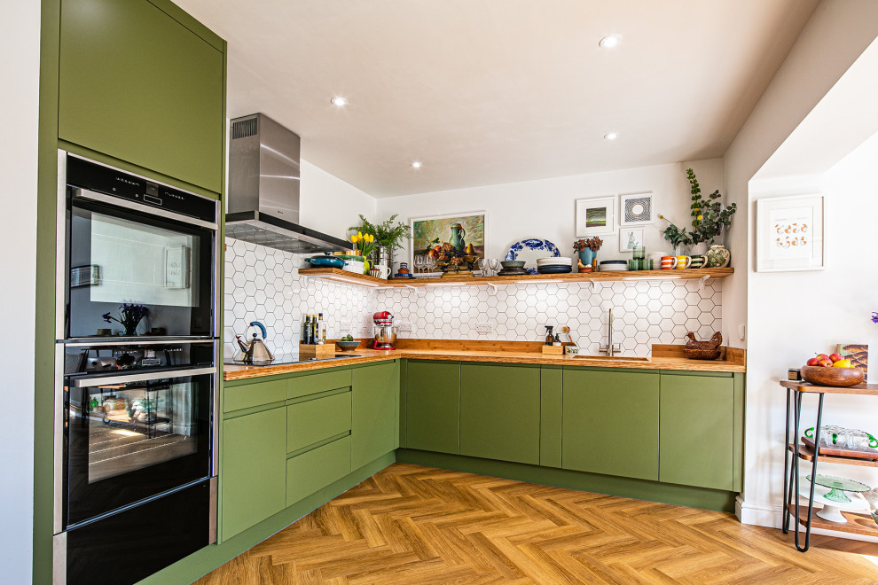 Mid-sized l-shaped open plan kitchen in Other with flat-panel cabinets, green cabinets, wood benchtops, white splashback, mosaic tile splashback, panelled appliances, medium hardwood floors, no island, brown floor and brown benchtop.