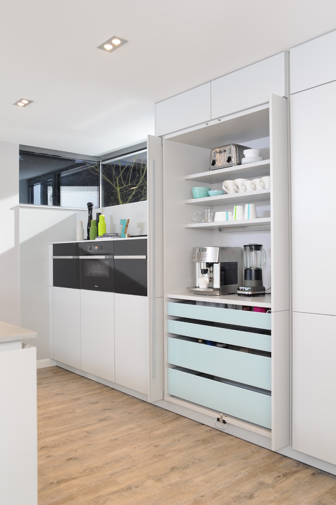 This is an example of an expansive contemporary galley open plan kitchen in Berlin with an integrated sink, flat-panel cabinets, white cabinets, concrete benchtops, white splashback, black appliances, light hardwood floors and with island.