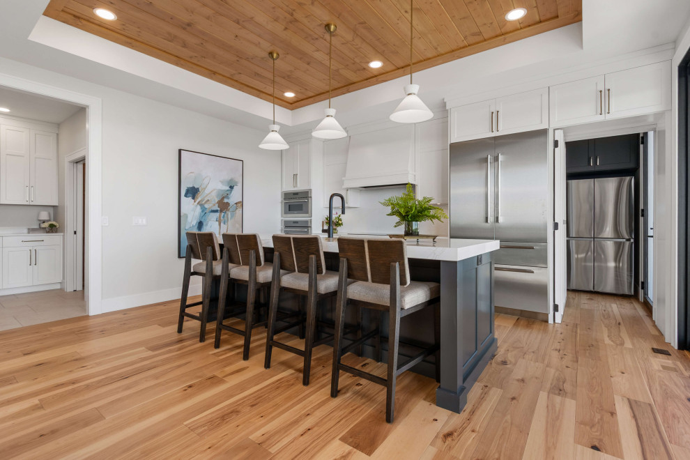 Transitional single-wall medium tone wood floor, brown floor and tray ceiling eat-in kitchen photo with an undermount sink, shaker cabinets, white cabinets, quartz countertops, white backsplash, quartz backsplash, stainless steel appliances, an island and white countertops