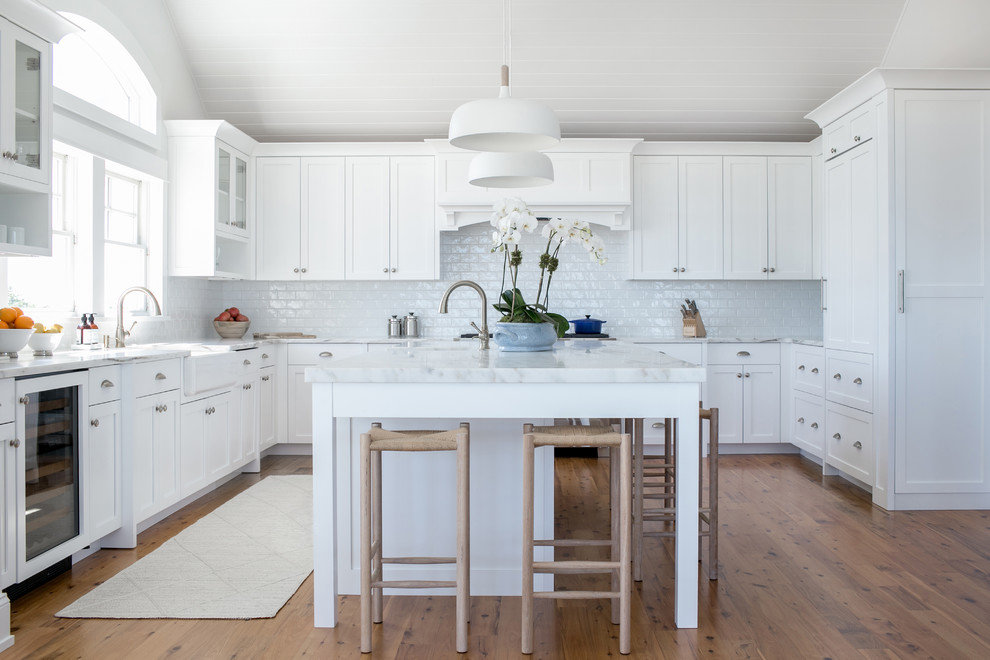 This is an example of a beach style u-shaped kitchen in New York with a farmhouse sink, shaker cabinets, white cabinets, white splashback, subway tile splashback, panelled appliances, medium hardwood floors, with island and brown floor.
