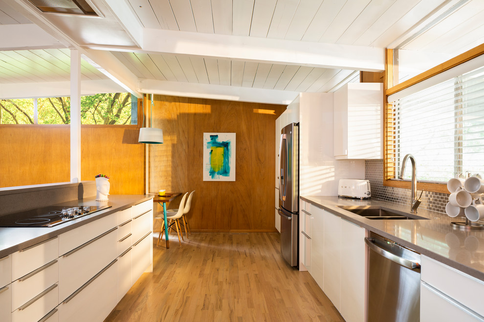 This is an example of a midcentury kitchen in Denver with a double-bowl sink, flat-panel cabinets, white cabinets, metallic splashback, metal splashback, stainless steel appliances, light hardwood floors and grey benchtop.