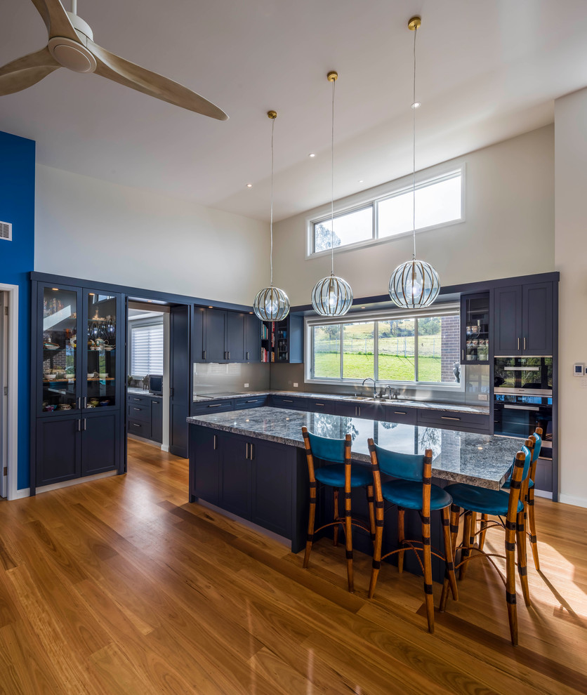 This is an example of an expansive contemporary kitchen in Sydney with a double-bowl sink, shaker cabinets, grey cabinets, granite benchtops, metallic splashback, glass sheet splashback, black appliances, light hardwood floors, with island, brown floor and grey benchtop.