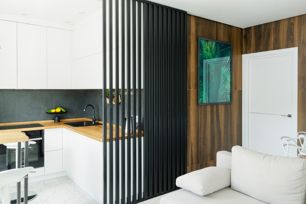 Small contemporary l-shaped open plan kitchen in Other with flat-panel cabinets, white cabinets, wood benchtops, black splashback, porcelain splashback, white floor, a drop-in sink, black appliances and brown benchtop.