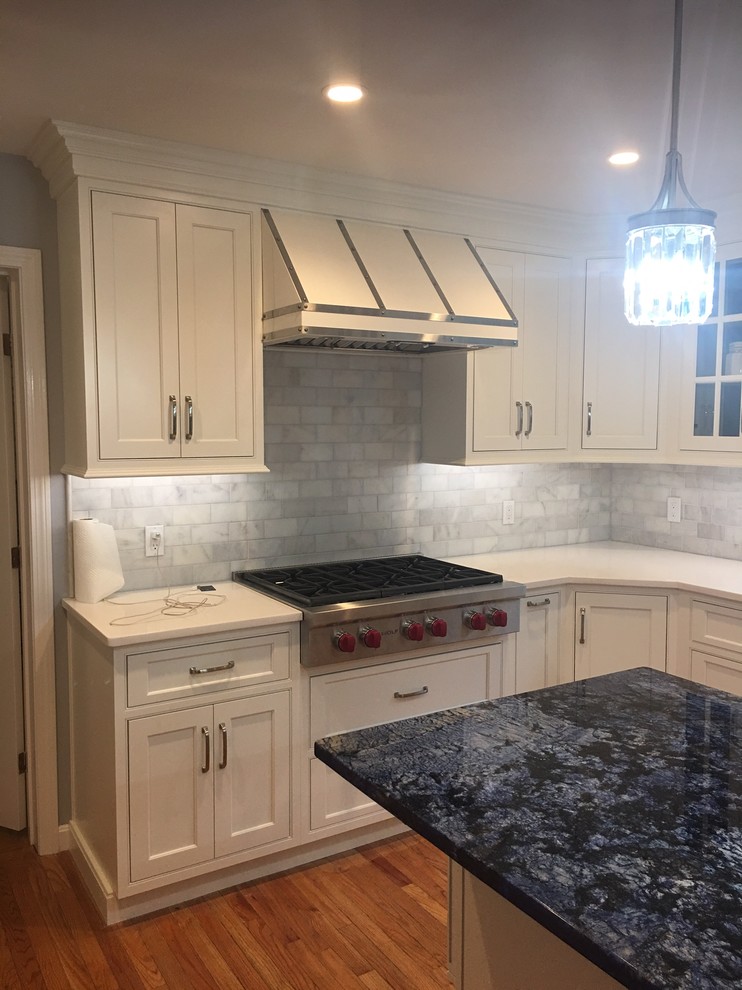 Mid-sized transitional l-shaped medium tone wood floor and brown floor kitchen photo in Boston with a farmhouse sink, shaker cabinets, white cabinets, granite countertops, gray backsplash, subway tile backsplash, stainless steel appliances and an island