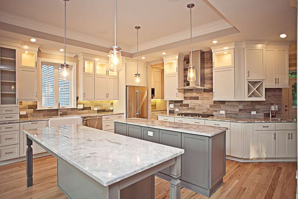 This is an example of a large transitional l-shaped open plan kitchen in New York with a farmhouse sink, recessed-panel cabinets, white cabinets, quartzite benchtops, multi-coloured splashback, stone tile splashback, stainless steel appliances, medium hardwood floors, multiple islands and brown floor.