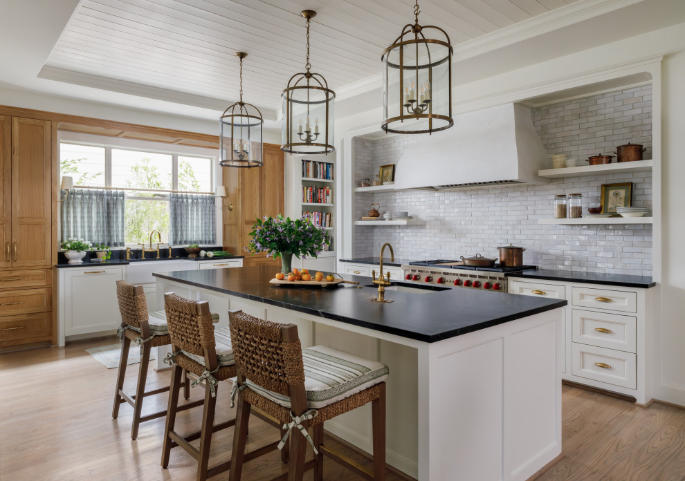 This is an example of a traditional eat-in kitchen in Houston with a farmhouse sink, recessed-panel cabinets, soapstone benchtops, white splashback, ceramic splashback, light hardwood floors, with island, brown floor, black benchtop and timber.