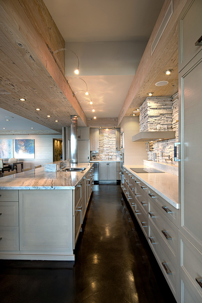 This is an example of a mid-sized transitional galley open plan kitchen in Miami with an undermount sink, recessed-panel cabinets, light wood cabinets, limestone benchtops, beige splashback, limestone splashback, stainless steel appliances, concrete floors, with island and brown floor.