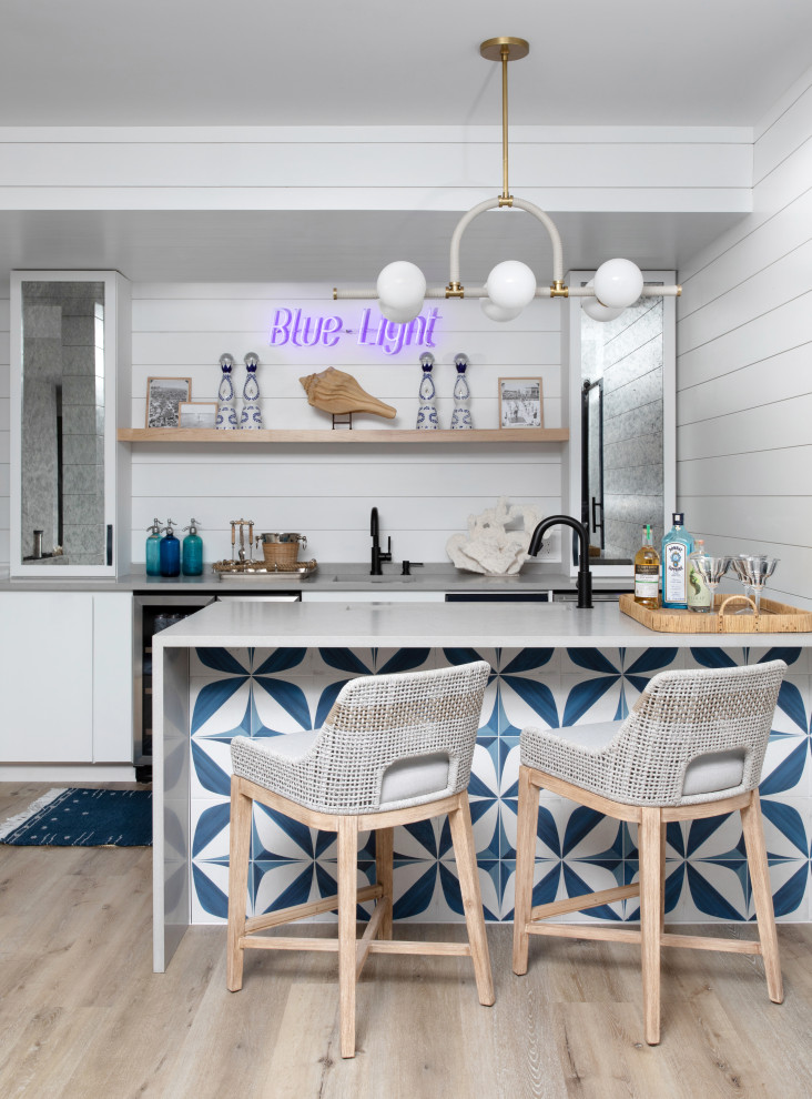 This is an example of an expansive beach style galley seated home bar in New York with an undermount sink, flat-panel cabinets, white cabinets, quartz benchtops, blue splashback, stone tile splashback, vinyl floors, brown floor and grey benchtop.