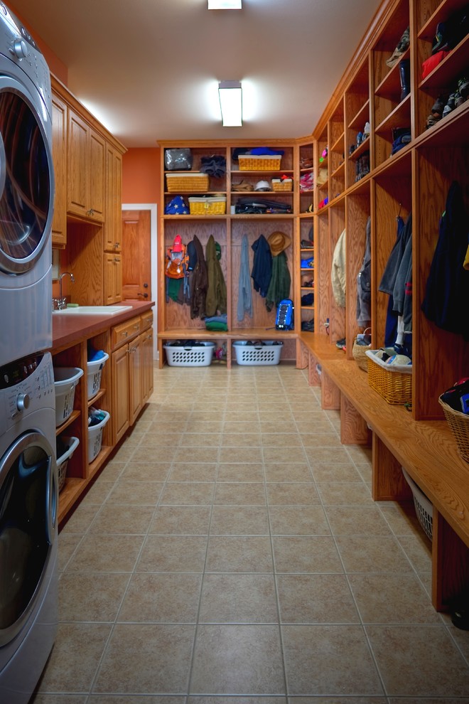 Inspiration for a mid-sized transitional galley utility room in Other with a drop-in sink, raised-panel cabinets, solid surface benchtops, orange walls, porcelain floors, a stacked washer and dryer and medium wood cabinets.