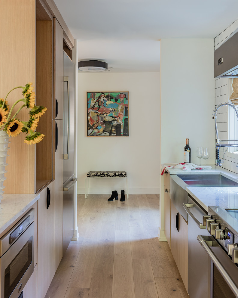 This is an example of a contemporary galley eat-in kitchen in Boston with a farmhouse sink, flat-panel cabinets, light wood cabinets, stainless steel appliances, white benchtop, light hardwood floors, white splashback, subway tile splashback, a peninsula and exposed beam.