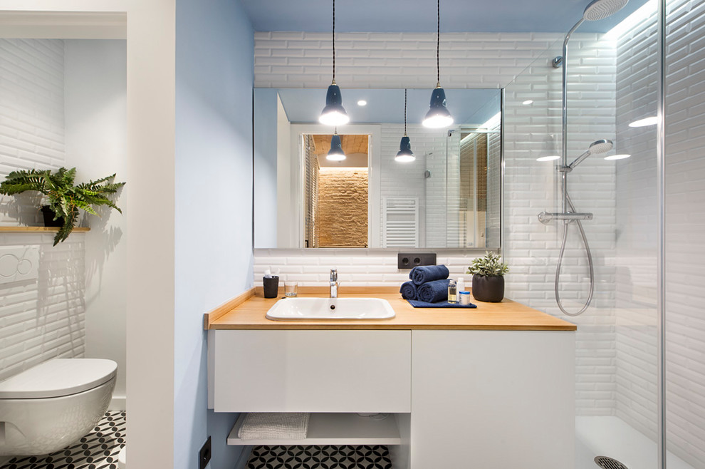 Photo of a mid-sized mediterranean 3/4 bathroom in Barcelona with flat-panel cabinets, white cabinets, a wall-mount toilet, white walls, ceramic floors, wood benchtops, multi-coloured floor, white tile, a drop-in sink and brown benchtops.