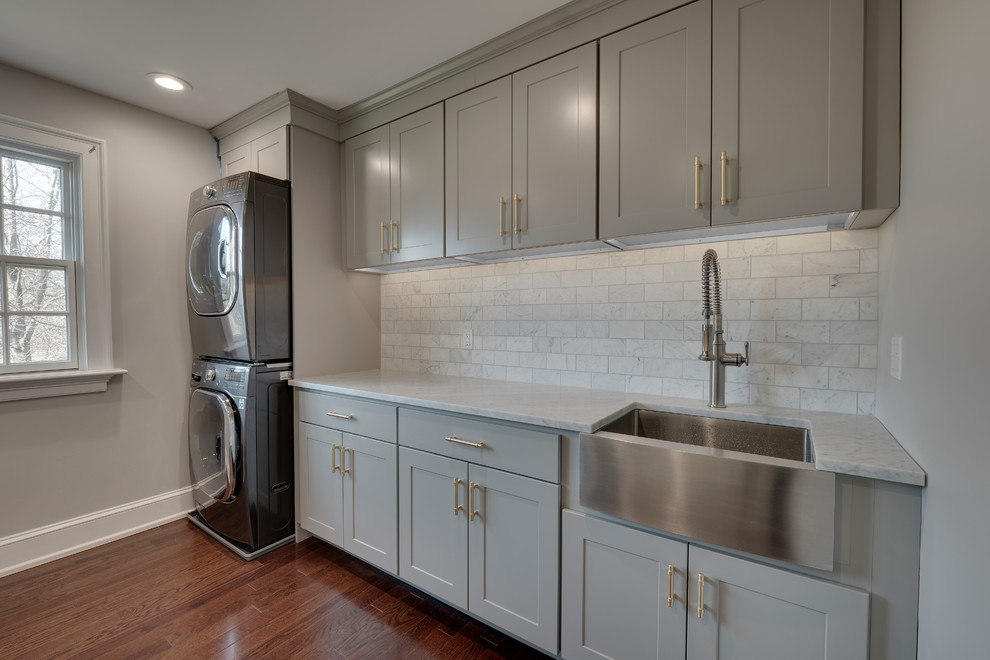 This is an example of a mid-sized transitional single-wall dedicated laundry room in Philadelphia with a farmhouse sink, recessed-panel cabinets, grey cabinets, marble benchtops, grey walls, medium hardwood floors, a stacked washer and dryer, brown floor and white benchtop.