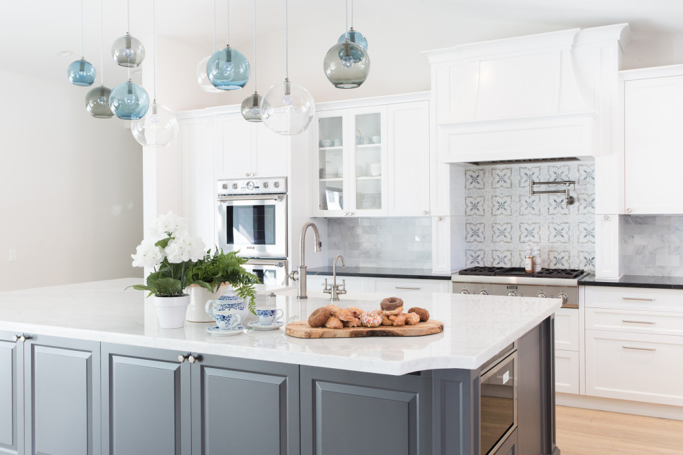 Transitional l-shaped open plan kitchen in Santa Barbara with a farmhouse sink, shaker cabinets, white cabinets, white splashback, marble splashback, stainless steel appliances, light hardwood floors, with island, beige floor, marble benchtops and black benchtop.