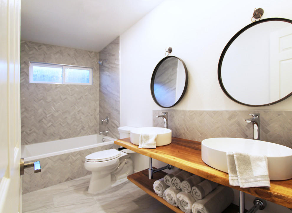 This is an example of a small kids bathroom in Other with open cabinets, medium wood cabinets, an alcove tub, a shower/bathtub combo, a two-piece toilet, gray tile, porcelain tile, white walls, porcelain floors, a vessel sink, wood benchtops, grey floor, brown benchtops, a double vanity and a floating vanity.