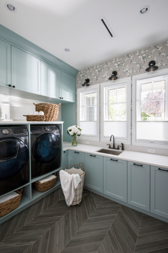 Transitional l-shaped laundry room in Vancouver with an undermount sink, shaker cabinets, blue cabinets, grey walls, a side-by-side washer and dryer, grey floor, white benchtop, wallpaper, quartz benchtops and porcelain floors.